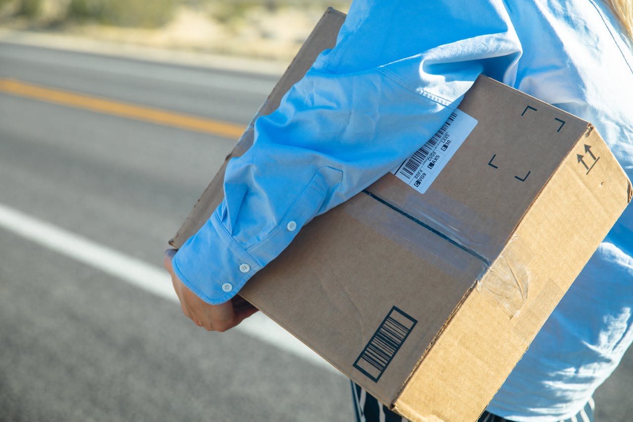 Close-up of a person carrying a cardboard box on a sunny road with a blue shirt.